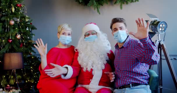 Alegre jovem casal vestindo máscaras faciais e chapéu de santa claus sentado em casa e acenando com as mãos — Vídeo de Stock