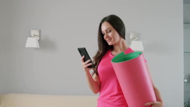 Joyeux jeune femme tient un tapis de yoga dans sa main et parle au téléphone. — Video