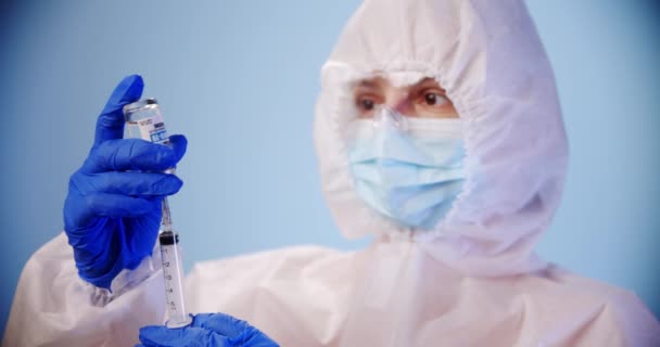 Verbleekte arts of verpleegkundige die persoonlijke beschermingsmiddelen draagt en het vaccin begint te vullen in de spuit met corona covid-19 vaccin in een laboratorium of ziekenhuis op een blauwe achtergrond. — Stockvideo