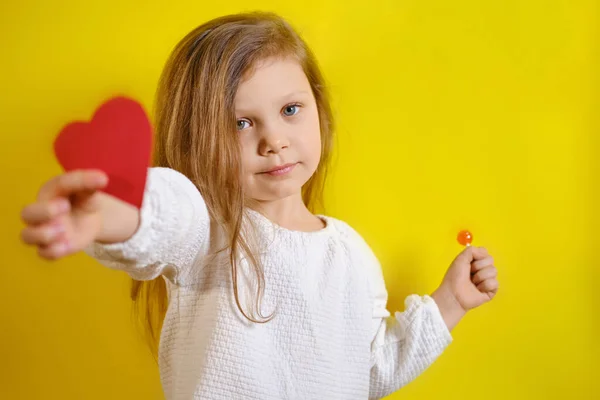 Retrato de niña sonriente sosteniendo un corazón rojo para ti y dulces en la segunda mano, aislado sobre fondo amarillo con espacio para copiar. — Foto de Stock