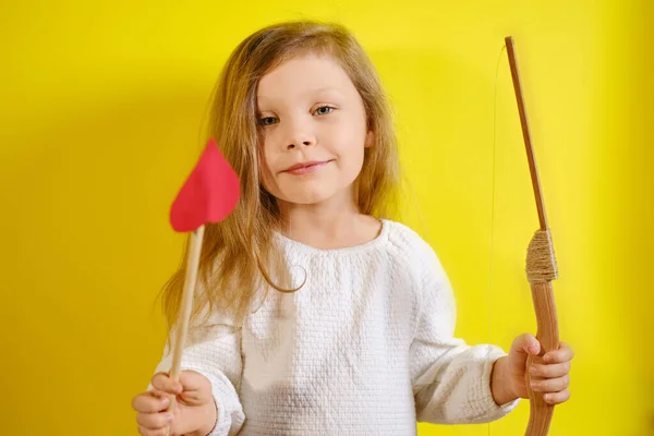 Retrato de niña sonriente sosteniendo una flecha en forma de corazón y una ballesta sobre fondo amarillo con espacio para copiar. — Foto de Stock