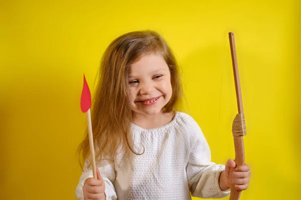 Retrato de niña feliz sonriente sosteniendo una flecha en forma de corazón y una ballesta sobre fondo amarillo con espacio para copiar. — Foto de Stock