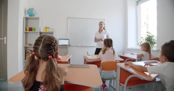 Iskolások ülnek asztaloknál az osztályteremben, aztán felemelik a kezüket, hogy válaszoljanak. Hátul. — Stock videók