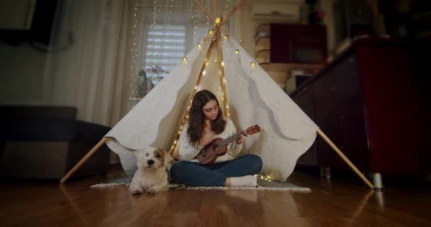 La chica se sienta en una tienda para niños con un perro Jack Russell, y toca la guitarra. Vercion 4 — Vídeo de stock