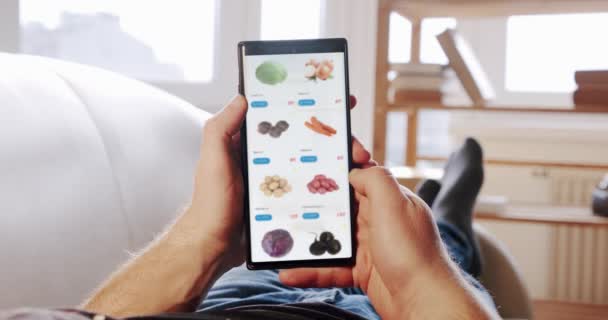 Man at Home Lying on Couch Orders Food In Online Store usando o aplicativo de entrega de alimentos em um smartphone. VPO — Vídeo de Stock