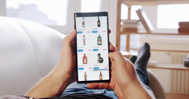 Человек дома лежит на диване и заказывает алкоголь в размытом интернет-магазине, используя приложение доставки еды на смартфоне. POV — стоковое видео