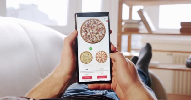 男人躺在家里沙发订单比萨饼在模糊的网上商店使用一个智能手机的递送应用程序。POV — 图库视频影像