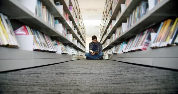 Estudiante universitario sentado en el piso de la biblioteca y leyendo un libro, entre filas de libros. — Vídeos de Stock