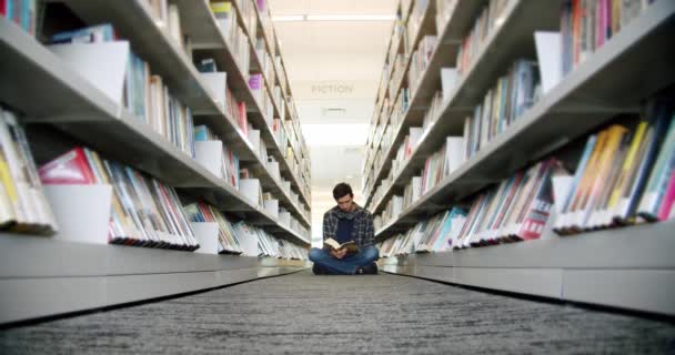 Студент колледжа сидит на полу в библиотеке и читает книгу, между рядами книг. Широкий обзор — стоковое видео