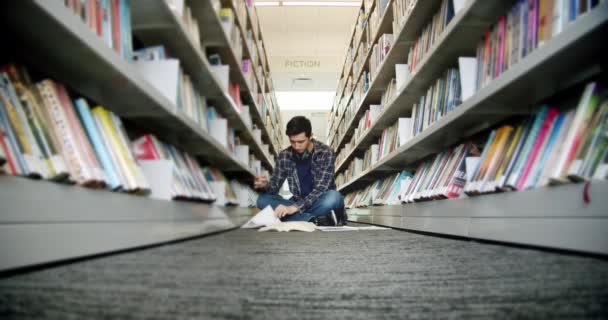 Széles kilátás egyetemista ül a padlón a könyvtárban, és olvasás egy könyvet, a sorok között könyvek. — Stock videók