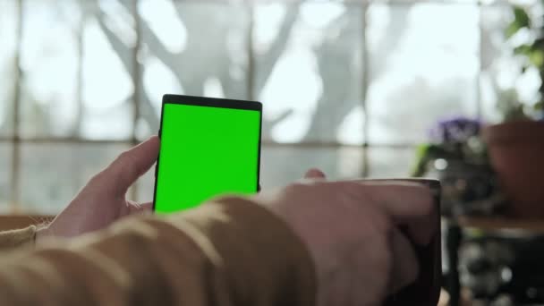 Una mano masculina sostiene un teléfono con una pantalla verde maqueta, y bebe café. De cerca. — Vídeos de Stock