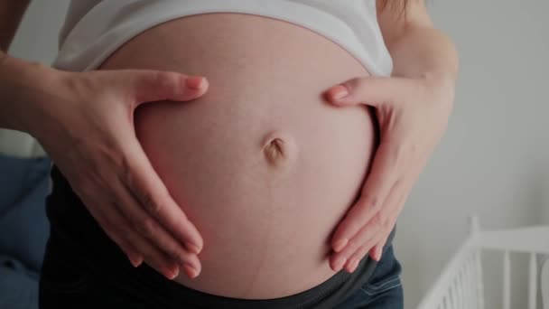 Крупный план молодой беременной женщины, гладящей большой живот на диване и придающей форму сердцу руками — стоковое видео