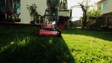Bir adam elektrikli çim biçme makinesiyle arka bahçedeki çimleri biçiyor. Yavaş çekim. Yakın çekim.