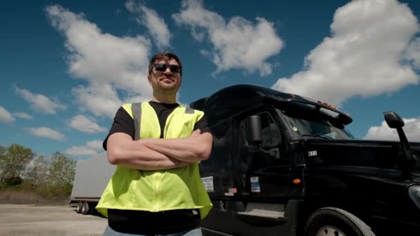 Sarı yelekli profesyonel kamyon şoförü kamyonetine yaklaşıyor ve kollarını kavuşturuyor. Arkasında da Kargo Karavanıyla birlikte Long Haul Semi-Truck park ediyor. — Stok video
