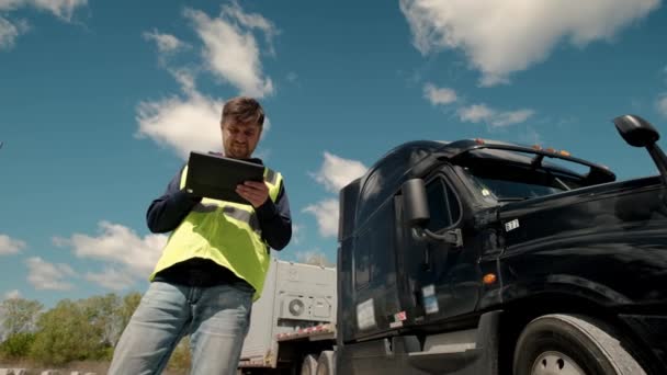 Kierowca ciężarówki stojący przy ciężarówce w żółtej kamizelce i używający tabletu do wypełnienia albumu. Szeroki strzał. — Wideo stockowe
