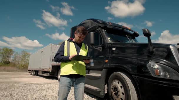 Truck driver standing by the truck in a yellow vest and using a tablet to fill a lookbook. Wide camera shot — Stock Video