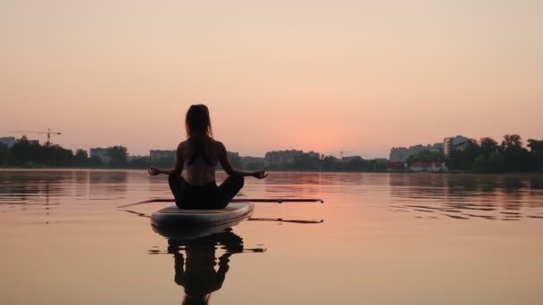 Mujer joven haciendo yoga en la tabla de SUP al atardecer. Vista en cámara lenta — Vídeo de stock