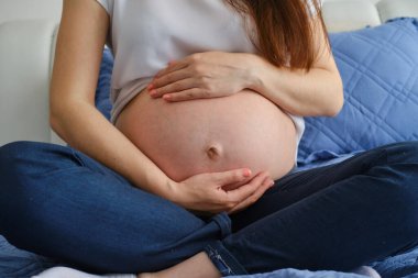 Genç hamile kadının yakın plan çekimi pencerenin önünde duruyor ve elleriyle karnına sarılıyor.