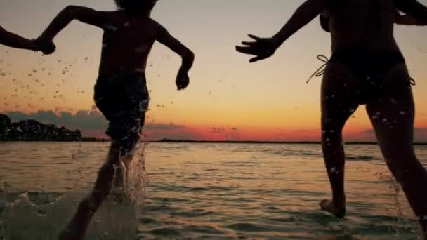 Silhouetten von Kindern, die bei Sonnenuntergang ins Wasser am Strand rennen. Glückliche, freundliche Familie. Zeitlupe — Stockvideo