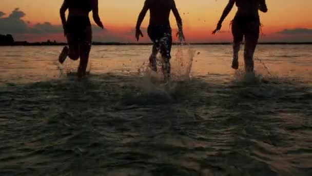 Napnyugtakor rohanó gyerekek sziluettjei a tengerparton a vízbe. Boldog barátságos családot. Lassú mozgás. — Stock videók