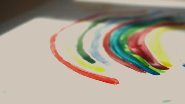 La petite fille tient un pinceau dans sa main et peint un arc-en-ciel avec des peintures colorées sur papier blanc. Macro shot — Video