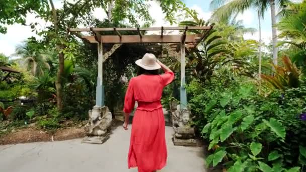 Молода брюнетка в червоній сукні гуляє по саду і обертається навколо, посміхаючись. Повільний рух. Широкий вигляд — стокове відео