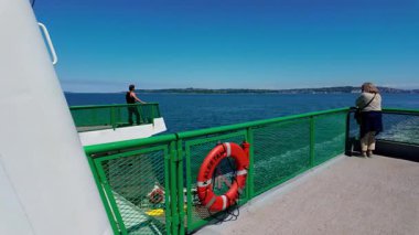 Yolcular feribotun açık güvertesinde dururlar. Yeşil korkuluklar ve parlak turuncu yaşam halkası deniz ortamını vurguluyor. ABD WA Seattle 2 Ağustos 2025. Yüksek kalite 4k görüntü