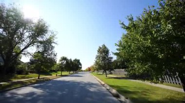POV güneşli bir yaz ya da sonbahar gününde sakin bir banliyö mahallesinde huzurlu bir şekilde araba sürer. Yeşil yapraklı ağaçlar caddeyi kaplıyor ve güneş ışığı sıcak ve davetkar bir atmosfer yaratıyor. Yüksek kalite 4k görüntü