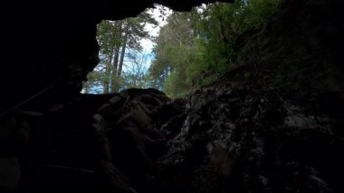 POV ormanı dar bir kaya boşluğundan geçiyor, mağaranın girişine benziyor. Büyük kayalar ve karanlık taş duvarlar sahneyi çevrelerken, güneş ışığı ve uzun ağaçlar ortaya çıkıyor. Yüksek kalite 4k görüntü