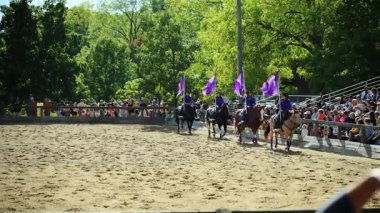 Binicilik performans takımı kumlu arenada ata biniyor. Sürücüler büyük mor bayraklar sallarken aynı anda senkronize oluş sergiliyorlar. USA IL Warrenville 2 Ekim 2025. Yüksek kalite 4k görüntü