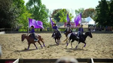 Binicilik performans takımı kumlu bir arenada ata biniyor. Sürücüler senkronize bir şekilde hareket ederken büyük mor bayraklar sallıyorlar. USA IL Warrenville 2 Ekim 2025. Yüksek kalite 4k görüntü