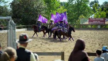 Binicilik performans takımı kumlu bir arenada ata biniyor. Sürücüler senkronize bir şekilde hareket ederken büyük mor bayraklar sallıyorlar. USA IL Warrenville 2 Ekim 2025. Yüksek kalite 4k görüntü