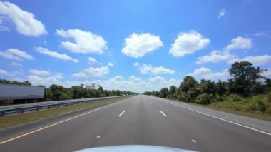 Güneşli bir günde açık bir otoyolda araba sürmek. Ağaçlar ve yeşillikler yol manzarasını oluştururken beyaz bulutlar parlak mavi bir gökyüzünde sürükleniyor. ABD FL Miami 02 Nisan 2025. Yüksek kalite 4k görüntü