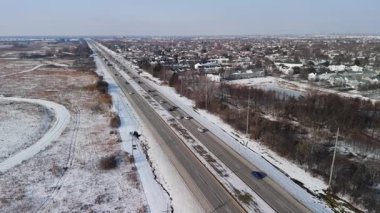 Kışın banliyö bölgesinde işlek bir otoyolun insansız hava aracı görüntüsü. Hafif kar yağışı çevredeki tarlaları, mahalleleri ve manzarayı kaplarken arabalar karla kaplı yollar boyunca seyahat ediyor. Yüksek kalite 4k görüntü