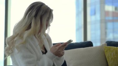 Yüksek katlı bir dairede modern bir kanepede oturan bir kadın, büyük pencereli ve arka planda şehir gökdelenleri olan telefonunu kontrol ediyor. Kapatın. Yüksek kalite 4k görüntü