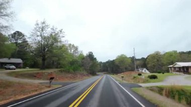 İlkbahar ağaçları ve yoğun yeşilliklerle çevrili yeni döşenmiş bir köy yolu boyunca sürüş görüntüsü. Virajlı yol, bulutlu gökyüzü ve sessiz orman manzarası. Yüksek kalite 4k görüntü