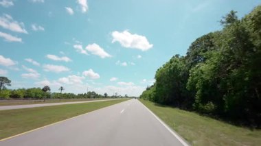 Açık mavi gökyüzünün altındaki bir bahar otobanı üzerindeki POV sürüş görüntüsü huzurlu bir kırsal alanda boş bir yol manzarası yakalıyor.