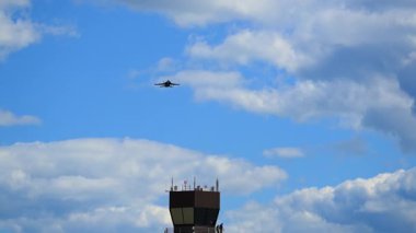 Askeri savaş uçağı, hava üssü altyapısı ile dinamik bir havacılık sahnesi yaratan mavi bulutlu bir gökyüzüne karşı havaalanı kontrol kulesinin yanından uçuyor. Alt manzara. Yüksek kalite 4k görüntü