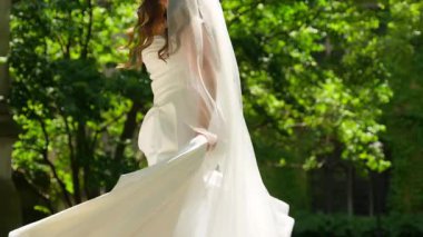 Beyaz bir gelinlik içindeki gelin, doğal güneş ışığında zarifçe döner, elbisesi ve duvağı hareket halindedir. Romantik açık hava düğün atmosferi neşeyi, zarafeti simgeliyor. Yüksek kalite 4k görüntü