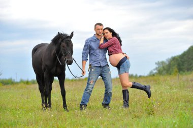 Çocuğunu bekliyor. Genç bir çift - uzun saçlı esmer, hamile; uzun boylu ve cesur, siyah atın dizginlerini tutan, çiftlikte bir yürüyüş.