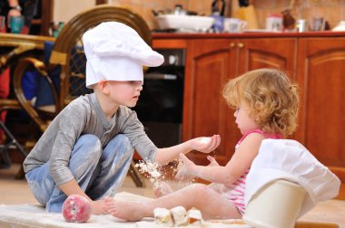 İki kardeş - erkek ve kız - şöminenin yanında aşçı şapkası takmış mutfakta un ile kirlenmiş, yemekle oynuyor, ortalığı dağıtıyor ve eğleniyorlar.