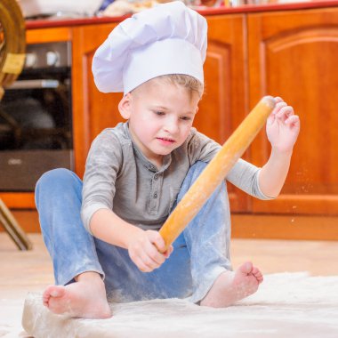 Çocuk mutfakta oturmuş un kirletiyor, yemekle oynuyor, ortalığı dağıtıyor ve eğleniyor.