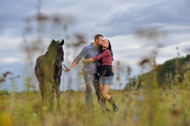 Sarılmak ve öpüşmek. Genç bir çift - uzun saçlı, uzun boylu, esmer, erkek uzun boylu, cesur, siyah atın dizginlerini tutan, çayırda bir yürüyüş. Arkalarında harika bir gökyüzü