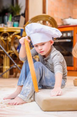 Çocuk mutfakta oturmuş un kirletiyor, yemekle oynuyor, ortalığı dağıtıyor ve eğleniyor.