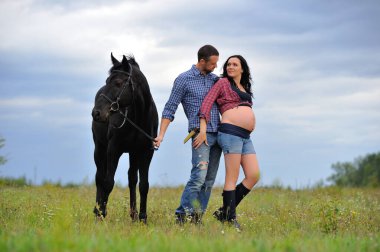 Çocuğunu bekliyor. Genç bir çift - uzun saçlı esmer, hamile; uzun boylu ve cesur, siyah atın dizginlerini tutan, çiftlikte bir yürüyüş.