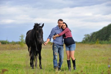 Çocuğunu bekliyor. Genç bir çift - uzun saçlı, uzun boylu, esmer, erkek uzun boylu, cesur, siyah atın dizginlerini tutan, çayırda bir yürüyüş. Arkalarında harika bir gökyüzü
