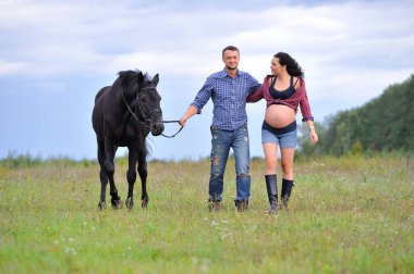 Çocuğunu bekliyor. Genç bir çift - uzun saçlı esmer, hamile; uzun boylu ve cesur, siyah atın dizginlerini tutan, çiftlikte bir yürüyüş.