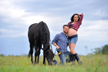 Çocuğunu bekliyor. Genç bir çift - uzun saçlı, uzun boylu, esmer, erkek uzun boylu, cesur, siyah atın dizginlerini tutan, çayırda bir yürüyüş. Arkalarında harika bir gökyüzü