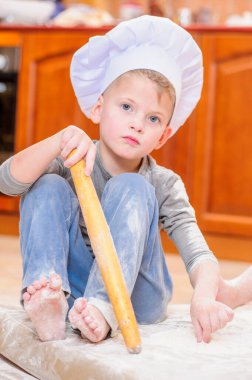 Çocuk mutfakta oturmuş un kirletiyor, yemekle oynuyor, ortalığı dağıtıyor ve eğleniyor.