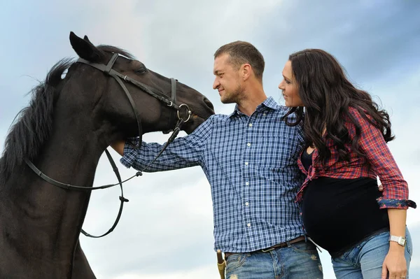 A walk on the ranch. Young couple - she is a brunette with long hair, pregnant; he is tall and brave, holding the reins of the black horse